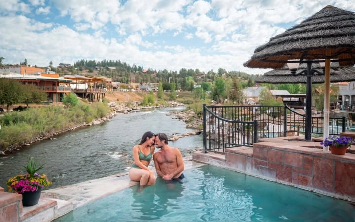 A man and woman kiss intimately in a blue pool, beside a river flowing past a town with buildings and forested hills under a cloudy sky.
