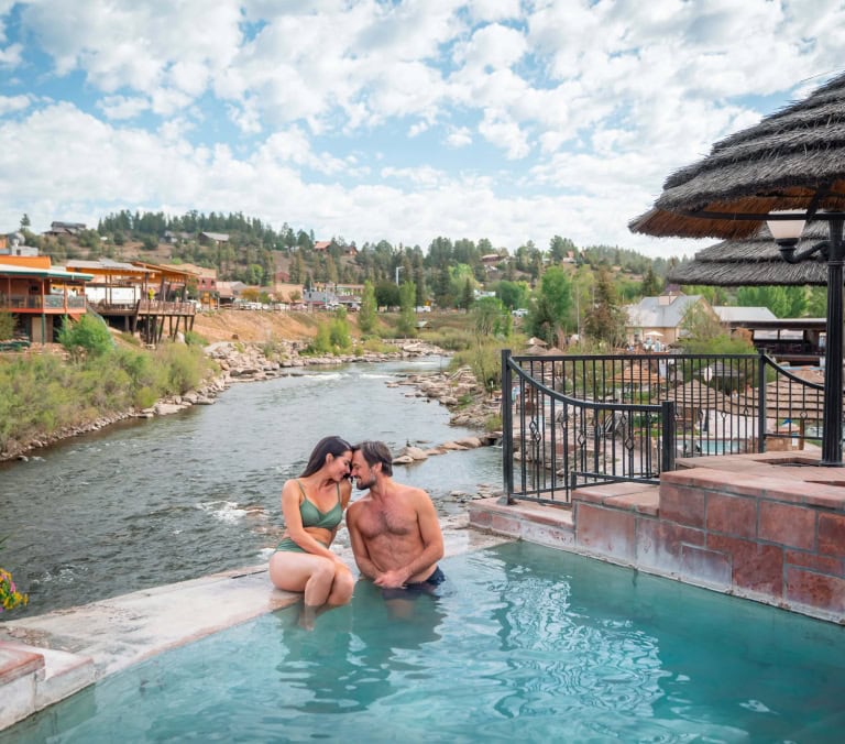 A man and woman kiss intimately in a blue pool, beside a river flowing past a town with buildings and forested hills under a cloudy sky.
