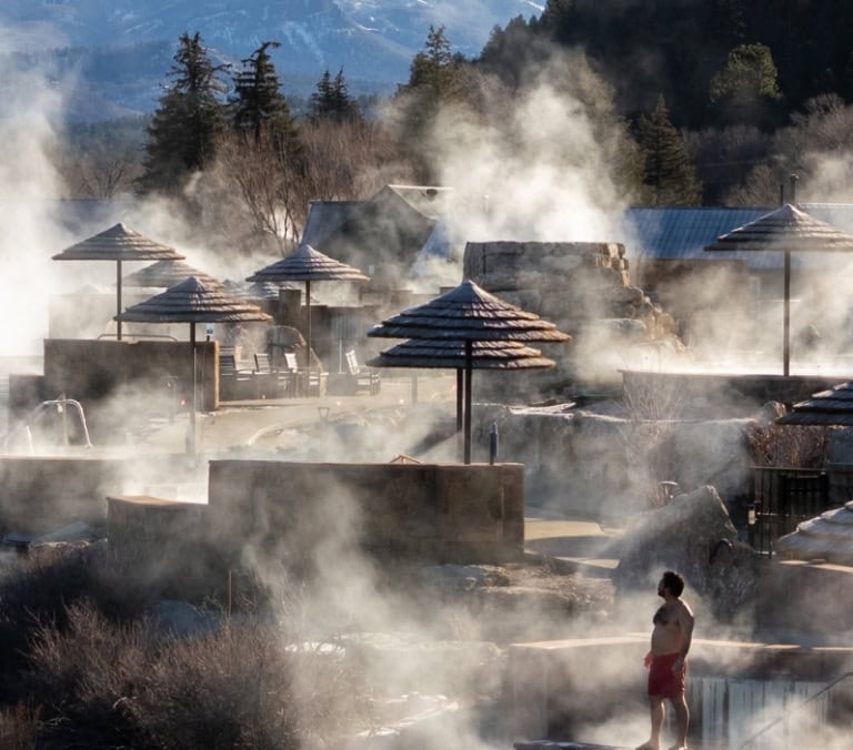 A multi-tiered hot springs complex steams heavily, featuring thatched umbrellas and a lone person, beside a river with a bridge, buildings, and snowy mountains beyond.
