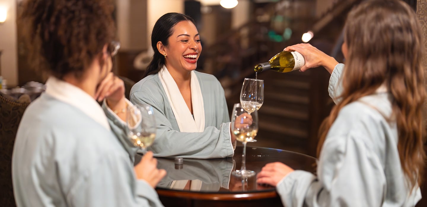 Three women in light blue robes share wine around a dark table. One laughs as another pours white wine from a bottle labeled 