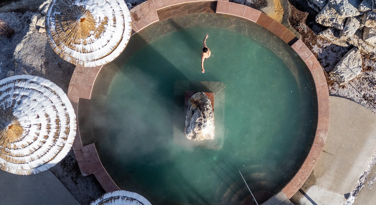 A person floats face-up in a steaming, circular hot spring pool featuring a large central rock. Snow-covered straw umbrellas and icy ground surround it.