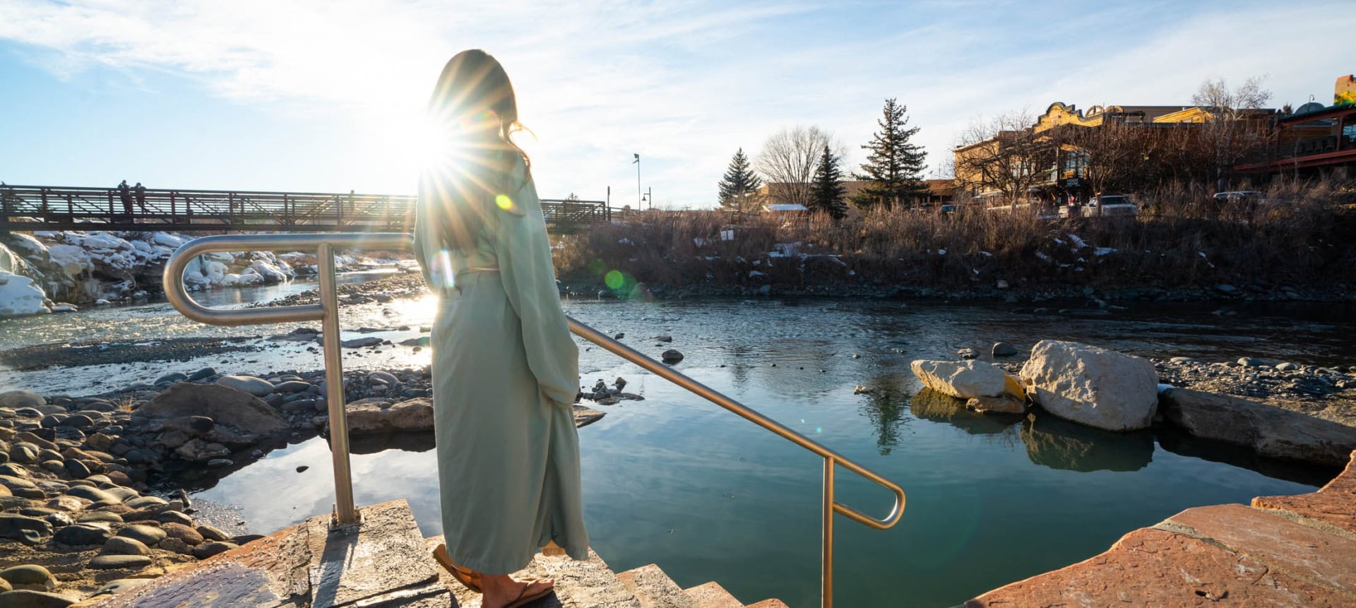 A person in a light robe stands on steps with a railing, looking at a hot spring adjacent to a river. A bridge, snowy banks, and buildings with a 