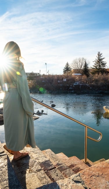 A person in a light robe stands on steps with a railing, looking at a hot spring adjacent to a river. A bridge, snowy banks, and buildings with a 