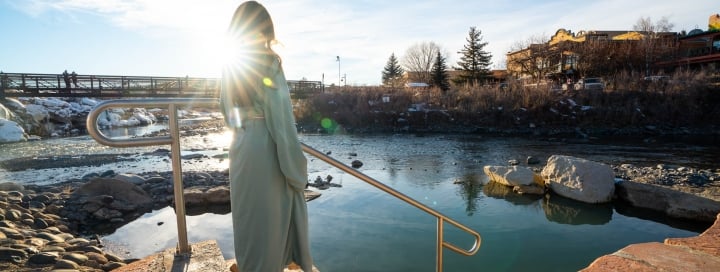 A person in a light robe stands on steps with a railing, looking at a hot spring adjacent to a river. A bridge, snowy banks, and buildings with a 