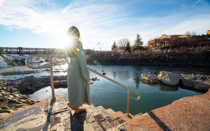 A person in a light robe stands on steps with a railing, looking at a hot spring adjacent to a river. A bridge, snowy banks, and buildings with a 