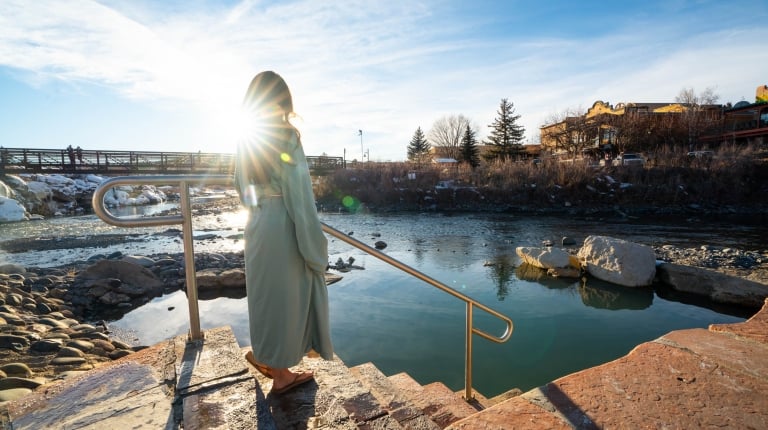 A person in a light robe stands on steps with a railing, looking at a hot spring adjacent to a river. A bridge, snowy banks, and buildings with a 