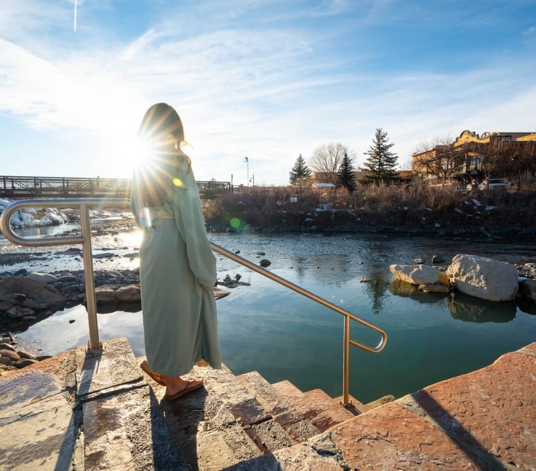 A person in a light robe stands on steps with a railing, looking at a hot spring adjacent to a river. A bridge, snowy banks, and buildings with a 