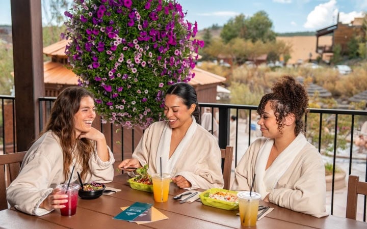 Three women in robes smile, eating salads and drinks at a patio table with a large purple flower basket. Text: SHARE THE SANCTUARY Share the experience with someone you love. SKINNY SPRINGS