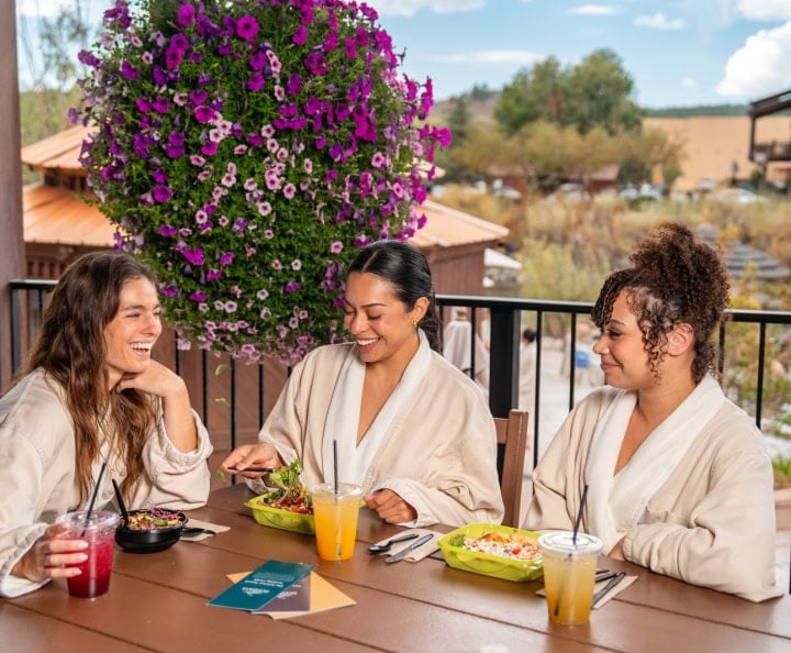 Three women in robes smile, eating salads and drinks at a patio table with a large purple flower basket. Text: SHARE THE SANCTUARY Share the experience with someone you love. SKINNY SPRINGS