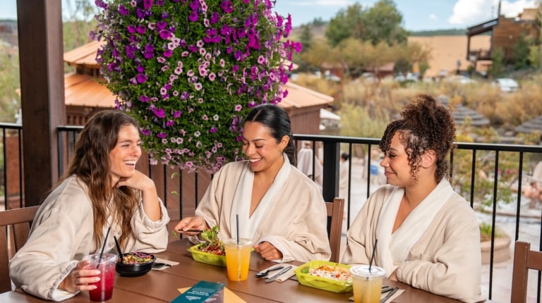 Three women in robes smile, eating salads and drinks at a patio table with a large purple flower basket. Text: SHARE THE SANCTUARY Share the experience with someone you love. SKINNY SPRINGS