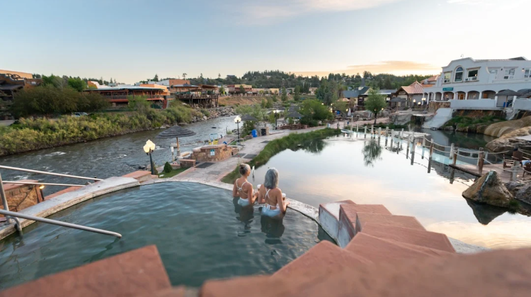 Two women sit in a hot spring pool, overlooking a river and resort town with other pools at dusk. Text: SPA