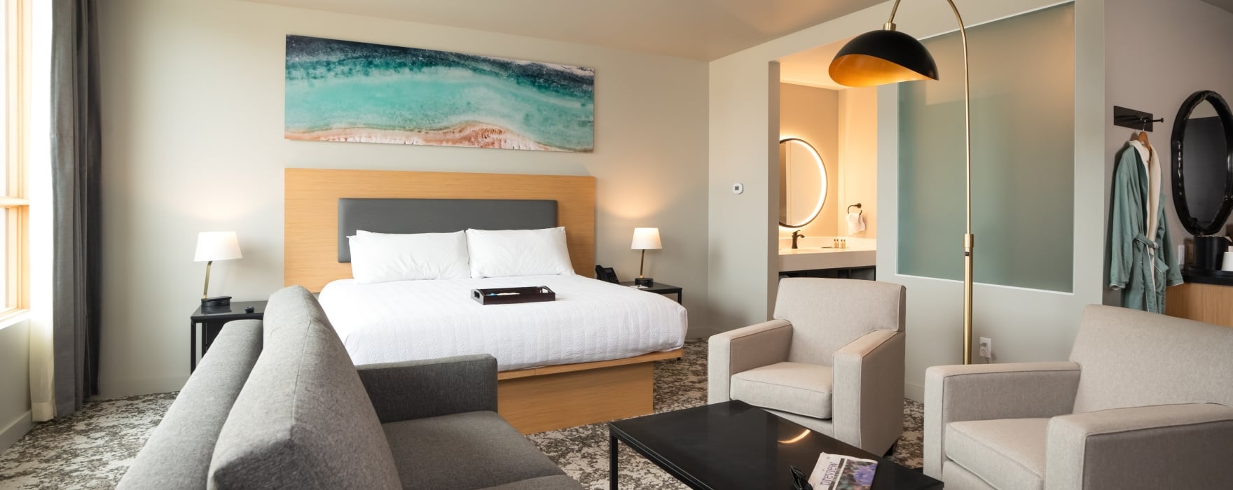Modern hotel room with a king bed, gray sofa, two armchairs, and coffee table holding 
