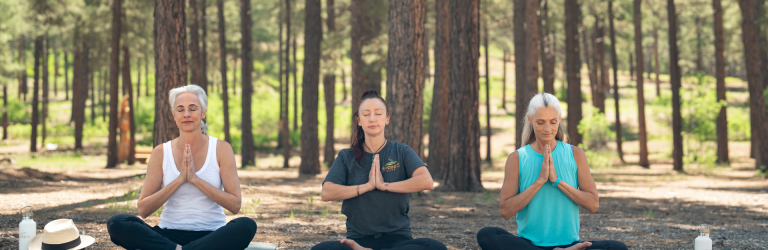 Three women sit cross-legged on yoga mats, meditating with closed eyes and hands in prayer position in a sunny pine forest. Text: SPRINGS, SPRINGS OUTDOORS, SPRINGS.