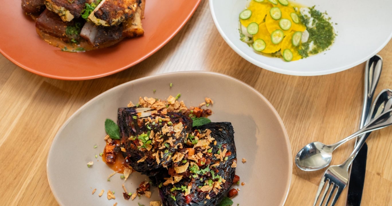 Three gourmet dishes are arranged on a light wooden table. Charred chicken, roasted purple cabbage, and yellow pasta with cucumber are presented on various plates and bowls, with cutlery.