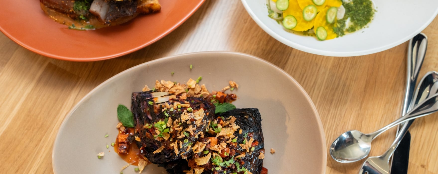 Three gourmet dishes are arranged on a light wooden table. Charred chicken, roasted purple cabbage, and yellow pasta with cucumber are presented on various plates and bowls, with cutlery.