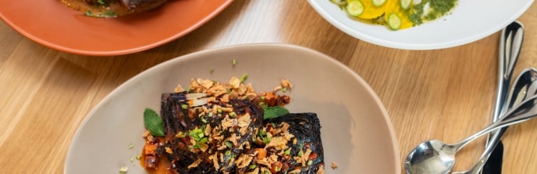 Three gourmet dishes are arranged on a light wooden table. Charred chicken, roasted purple cabbage, and yellow pasta with cucumber are presented on various plates and bowls, with cutlery.
