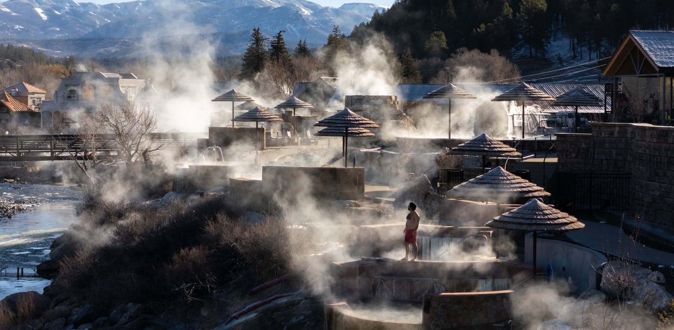 A person in red shorts stands by steaming terraced hot spring pools with thatched umbrellas, a river, and distant snow-capped mountains and evergreen trees in the background.