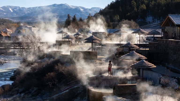 A person in red shorts stands by steaming terraced hot spring pools with thatched umbrellas, a river, and distant snow-capped mountains and evergreen trees in the background.