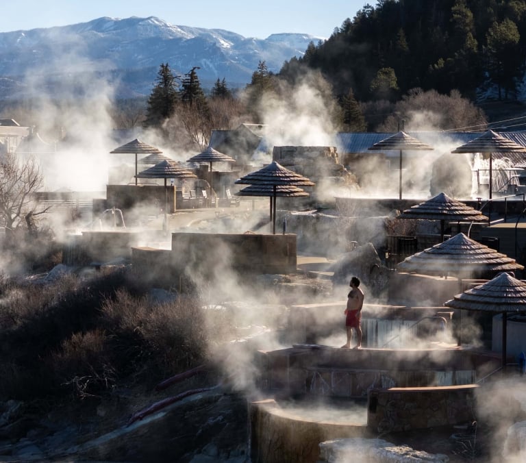 A person in red shorts stands by steaming terraced hot spring pools with thatched umbrellas, a river, and distant snow-capped mountains and evergreen trees in the background.