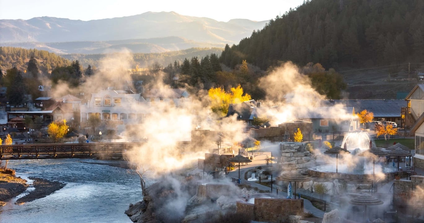 Numerous outdoor hot springs release steam around buildings. A river flows through a mountain town with autumn foliage, under warm, hazy light. Text: THE SPRINGS