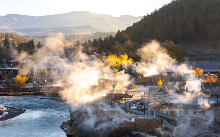 Numerous outdoor hot springs release steam around buildings. A river flows through a mountain town with autumn foliage, under warm, hazy light. Text: THE SPRINGS
