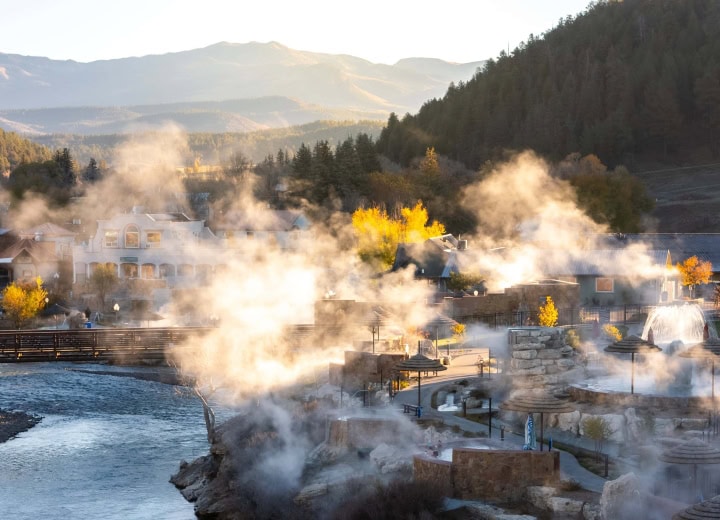 Numerous outdoor hot springs release steam around buildings. A river flows through a mountain town with autumn foliage, under warm, hazy light. Text: THE SPRINGS