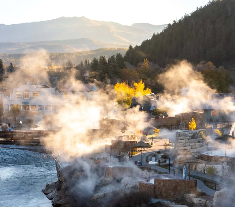 Numerous outdoor hot springs release steam around buildings. A river flows through a mountain town with autumn foliage, under warm, hazy light. Text: THE SPRINGS