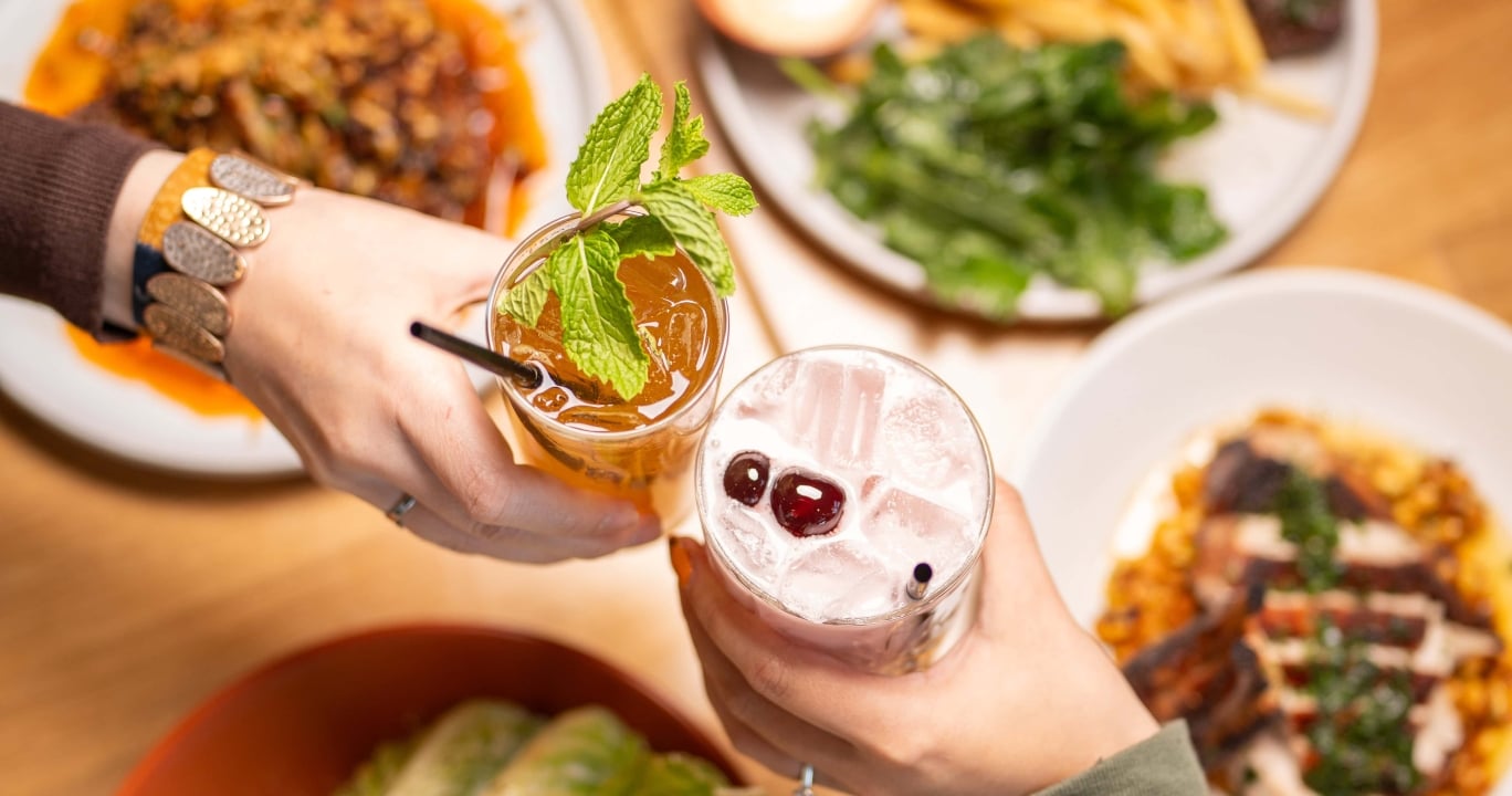 Two hands clink cocktails above a wooden table filled with plates of food including fries, salad, and grilled items, viewed from above.