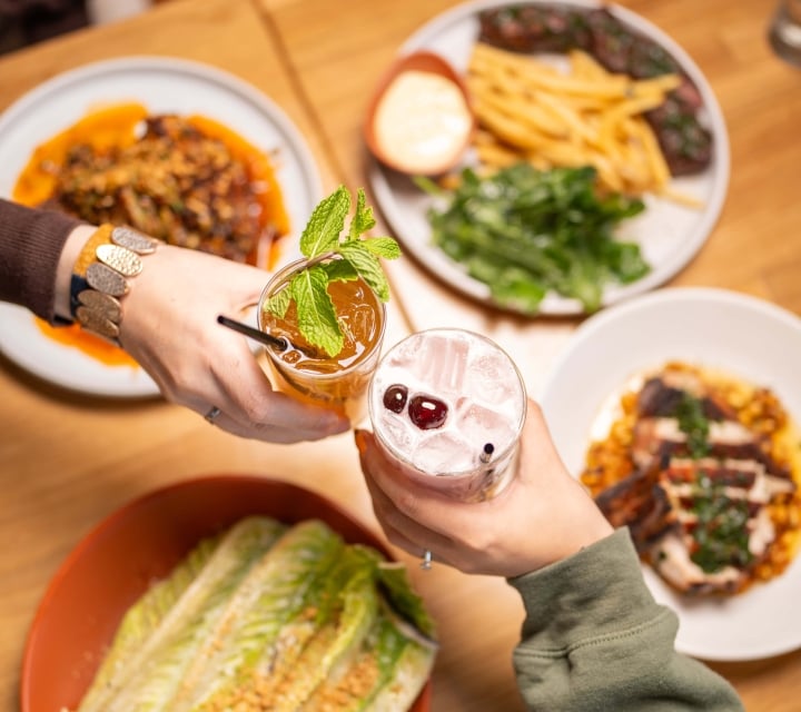 Two hands clink cocktails above a wooden table filled with plates of food including fries, salad, and grilled items, viewed from above.