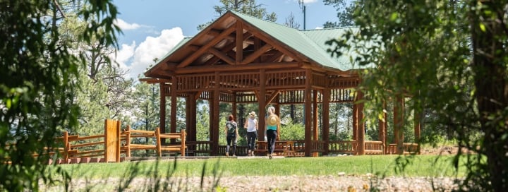 Three women walk past a large wooden gazebo with a green roof on green grass, surrounded by pine trees under a blue, cloudy sky.