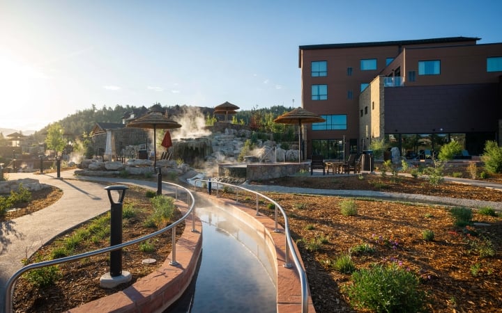 Steaming river-like hot spring curves through a resort, surrounded by paved paths, rock formations with waterfalls, straw umbrellas, and brown multi-story buildings under a clear sky.