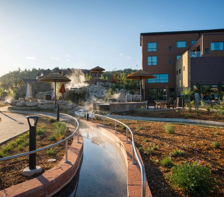 Steaming river-like hot spring curves through a resort, surrounded by paved paths, rock formations with waterfalls, straw umbrellas, and brown multi-story buildings under a clear sky.