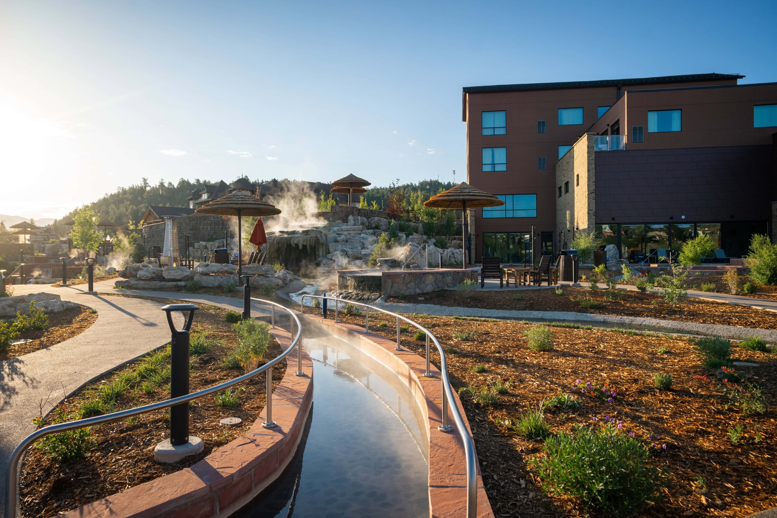 Steaming river-like hot spring curves through a resort, surrounded by paved paths, rock formations with waterfalls, straw umbrellas, and brown multi-story buildings under a clear sky.