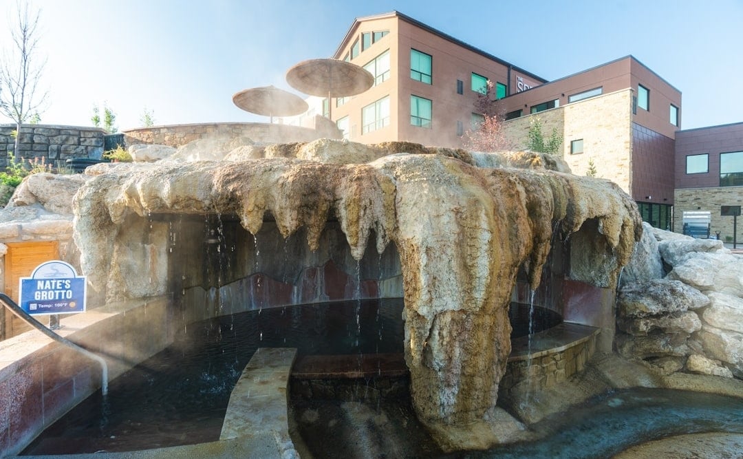 NATE'S GROTTO (Temp: 100°F ♨) is a rock grotto with water cascading into a steaming pool, set outdoors with a modern building and umbrellas in the background.