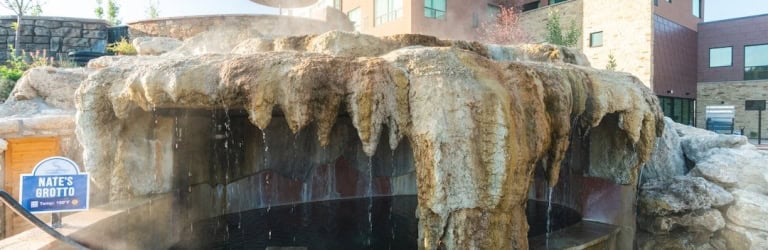 NATE'S GROTTO (Temp: 100°F ♨) is a rock grotto with water cascading into a steaming pool, set outdoors with a modern building and umbrellas in the background.