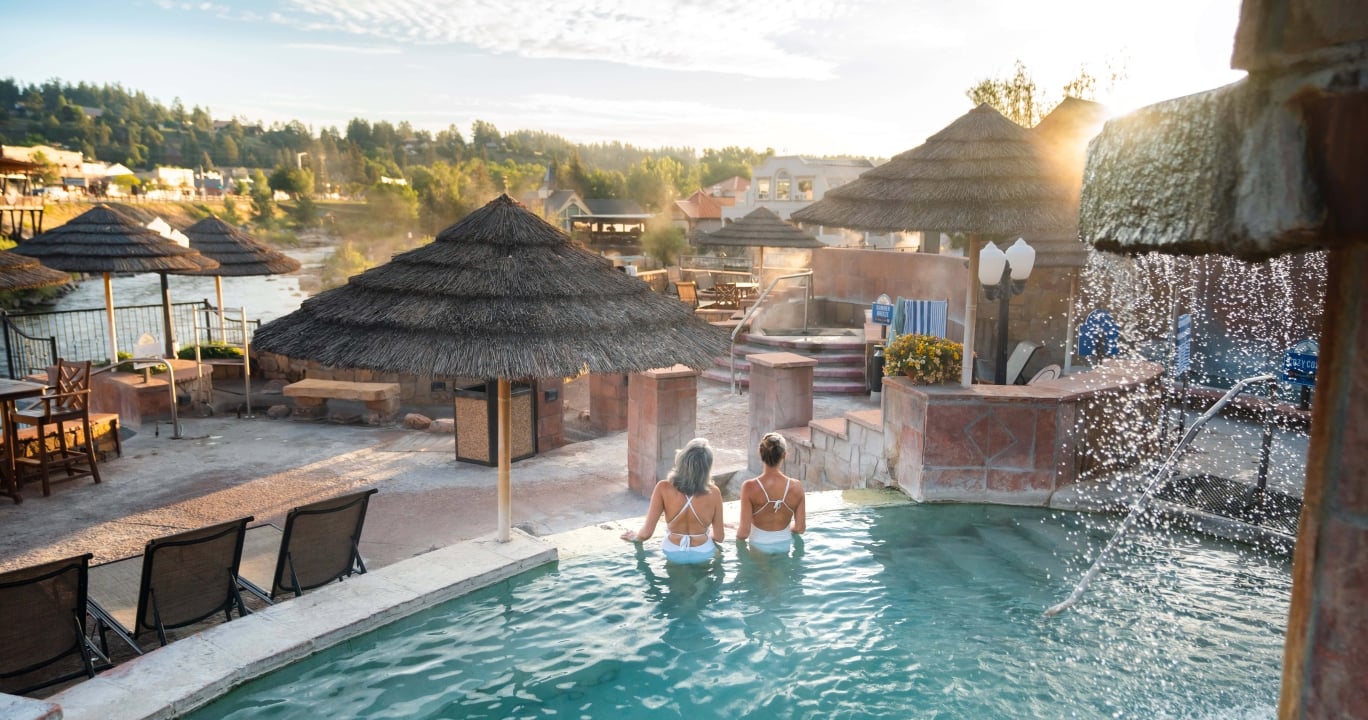 Two women face away, sitting in a turquoise outdoor spa pool. Thatch umbrellas, a river, and a town on a hill frame the scene. Text: SUMMER BREEZE.
