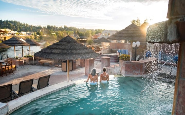 Two women face away, sitting in a turquoise outdoor spa pool. Thatch umbrellas, a river, and a town on a hill frame the scene. Text: SUMMER BREEZE.