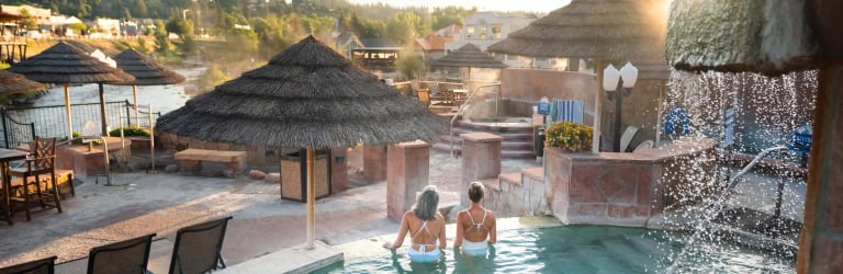 Two women face away, sitting in a turquoise outdoor spa pool. Thatch umbrellas, a river, and a town on a hill frame the scene. Text: SUMMER BREEZE.