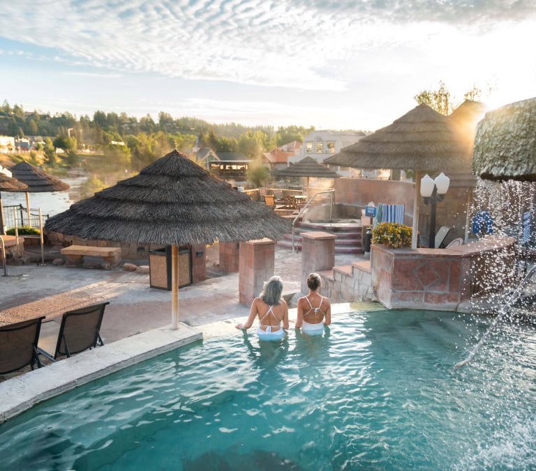 Two women face away, sitting in a turquoise outdoor spa pool. Thatch umbrellas, a river, and a town on a hill frame the scene. Text: SUMMER BREEZE.