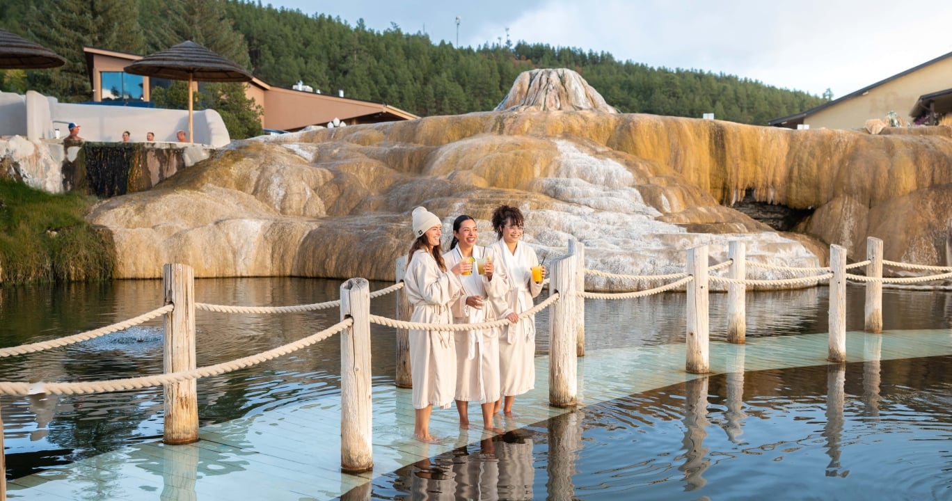 Three women in robes stand barefoot on a platform, holding drinks and smiling in a hot spring pool. Tiered orange mineral formations rise behind them, with trees and buildings in the distance.