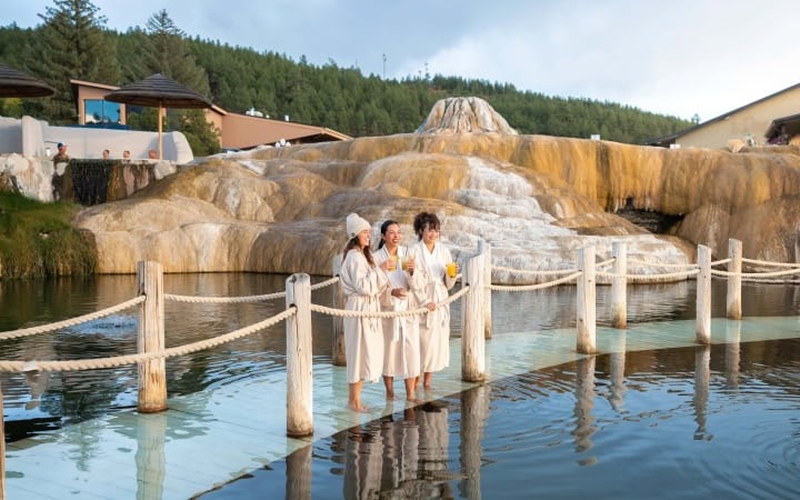 Three women in robes stand barefoot on a platform, holding drinks and smiling in a hot spring pool. Tiered orange mineral formations rise behind them, with trees and buildings in the distance.