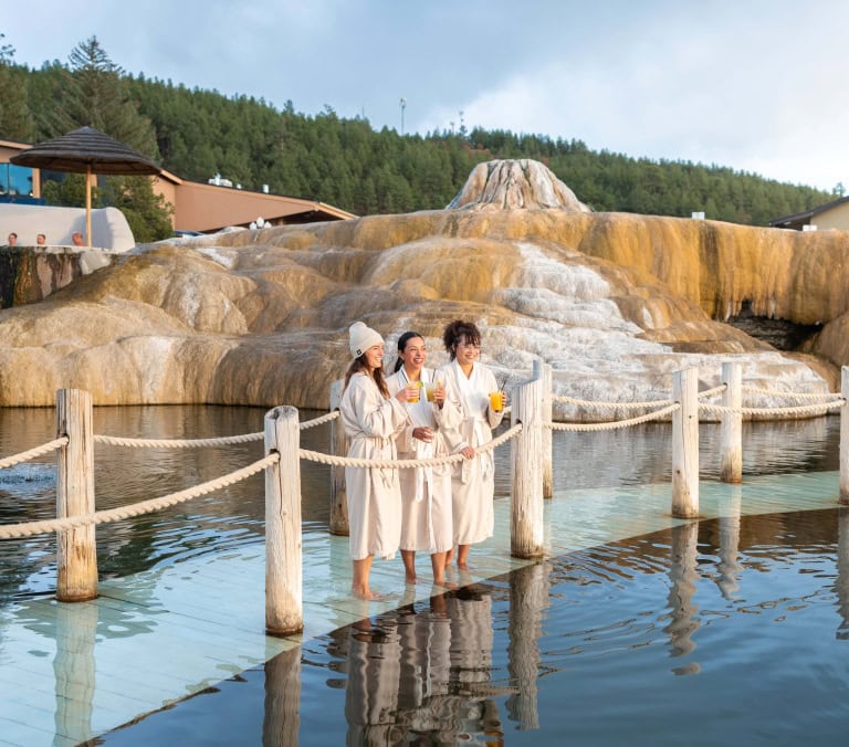 Three women in robes stand barefoot on a platform, holding drinks and smiling in a hot spring pool. Tiered orange mineral formations rise behind them, with trees and buildings in the distance.