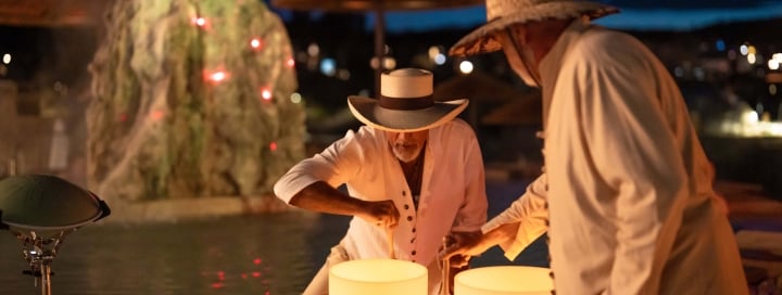 Two people in straw hats play glowing crystal singing bowls on a wooden table, at night by a pool with a lighted rock and distant town lights.