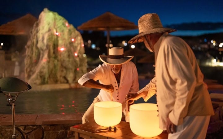 Two people in straw hats play glowing crystal singing bowls on a wooden table, at night by a pool with a lighted rock and distant town lights.
