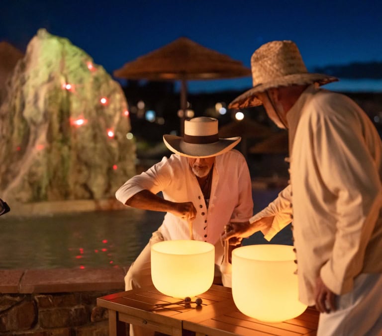 Two people in straw hats play glowing crystal singing bowls on a wooden table, at night by a pool with a lighted rock and distant town lights.