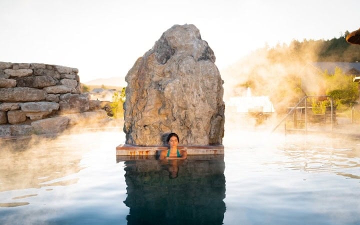 A woman relaxes in a steaming hot spring pool, eyes closed, against a large rock formation and stone wall, surrounded by rising mist under a bright sky.
