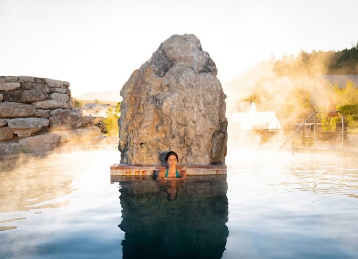 A woman relaxes in a steaming hot spring pool, eyes closed, against a large rock formation and stone wall, surrounded by rising mist under a bright sky.