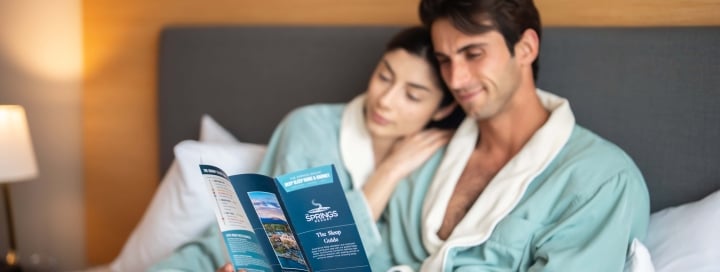 A man and woman in bathrobes read a brochure together while relaxing on a bed in a bright hotel room.