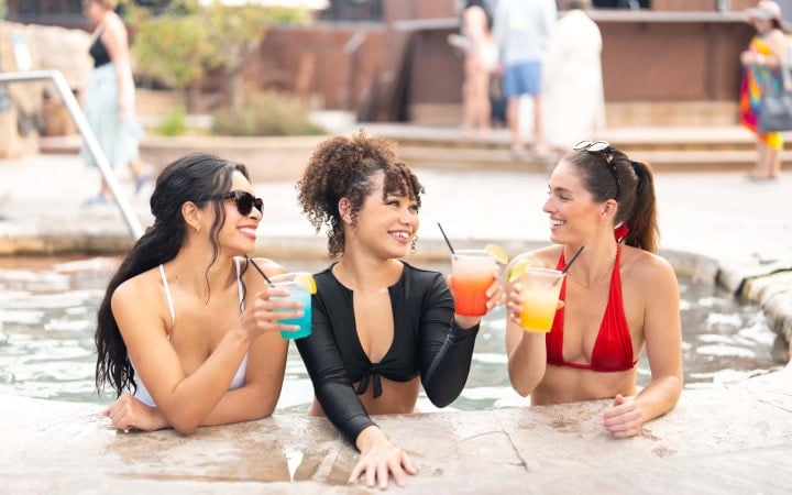 Three women, smiling, hold colorful cocktails (blue, orange, yellow) while socializing in a pool, leaning on its wet edge. Others are visible in the blurred background.