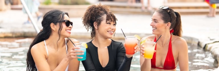 Three women, smiling, hold colorful cocktails (blue, orange, yellow) while socializing in a pool, leaning on its wet edge. Others are visible in the blurred background.